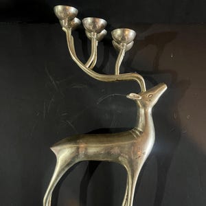 May include: Silver-coloured deer-shaped candle holder with four candle cups. The deer has a slender body and long legs, with the candle cups branching out from its antlers. The metal has a slightly aged appearance.