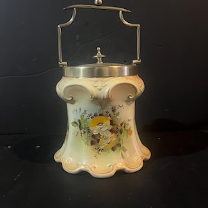 Könnte beinhalten: Ein cremefarbener Keksbehälter aus Keramik mit einem silberfarbenen Metallgriff und Deckel. Die Dose hat ein Blumendesign mit gelben und lila Blumen. Der Griff ist gebogen und der Deckel hat einen kleinen Knauf.