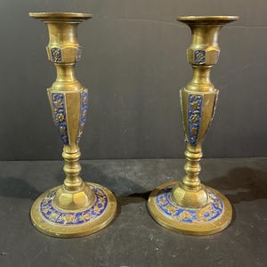 Könnte beinhalten: Ein Paar antike Messing-Kerzenständer mit blauen und goldenen Emaille-Details. Die Kerzenständer haben einen runden Sockel, einen schlanken Stiel und eine ausgestellte Oberseite. Das blaue Emaille zeigt florale Muster.