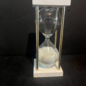 Puede incluir: Un reloj de arena con un bulbo de vidrio transparente lleno de arena blanca, sostenido entre dos bases cuadradas de madera blanca. Cuatro varillas metálicas delgadas de tono dorado conectan las piezas de madera, creando un marco vertical.