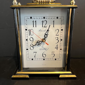 May include: A rectangular Brentwood quartz mantel clock featuring a white face with black numerals, ornate black hands, and a thin red second hand. The clock is encased in a black frame with gold-tone metal accents, a top handle, and two decorative finials.