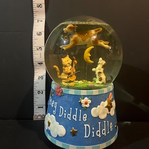 May include: A whimsical snow globe featuring a brown and white cow jumping over a crescent moon, a cat playing a violin, and a dog. The base is blue with the words "Hey Diddle Diddle" and decorative clouds, stars, and flowers.