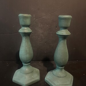 Peut inclure: Une paire de bougeoirs vert sauge dotés d'un pied tourné de style balustre et d'une base hexagonale. La surface de chaque bougeoir présente un grain de bois texturé visible avec des variations subtiles dans la couleur verte mate.