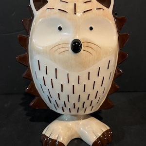 May include: A ceramic hedgehog figurine featuring a cream-coloured face with a black nose and eyes, accented by brown painted quills along the outer edge. The body displays a pattern of small brown dashes, and the figure stands on two feet.