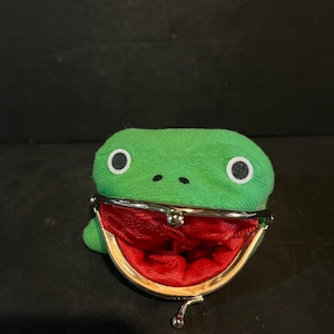 Peut inclure: Un porte-monnaie en tissu vert en forme de tête de grenouille avec de grands yeux blancs et noirs. Il est doté d'un fermoir à cliquet en métal argenté qui s'ouvre pour révéler une doublure intérieure en tissu rouge vif, créant une bouche ouverte.