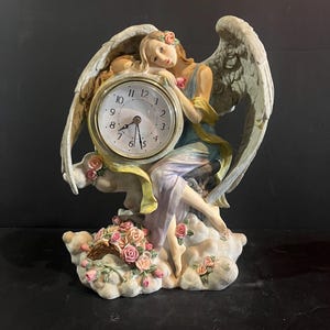 May include: A decorative angel clock figurine. The angel is seated on a cloud, with large feathered wings and a floral crown. The clock face is white with black numerals and gold trim. A bouquet of pink roses sits at the base.