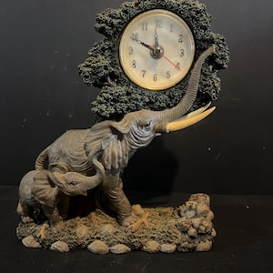 Può includere: Un orologio decorativo con una famiglia di elefanti e un uccello. Il quadrante dell'orologio è circondato da una cornice strutturata verde scuro. Gli elefanti sono grigi con zanne d'avorio e l'uccello è appollaiato sopra.