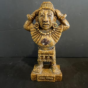 Op de afbeelding: Een getextureerd, goudbruin harsbeeldje van een figuur met de handen omhoog naar het hoofd. De figuur draagt een brede, geplooide kraag, ronde oorbellen en een rok met hangende kwastjes. Het staat op een rechthoekige voet met een klein plaatje.