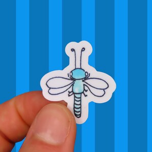 May include: A small die-cut sticker featuring a hand-drawn illustration of a dragonfly. The insect has a light blue watercolor-style body, dark blue outlines, two pairs of wings, and two antennae, all set against a white background border.