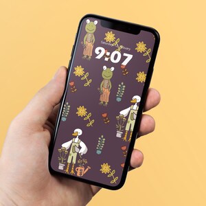 May include: A smartphone displaying a dark purple wallpaper featuring a repeating pattern of illustrated frogs and ducks wearing overalls, surrounded by yellow flowers and potted plants. The screen shows the time 9:07 and Saturday, 12 February.