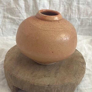May include: A small, round, peach-colored ceramic vase with a narrow opening. The vase sits on a rustic, round wooden stand. The vase has a textured surface and a slightly darker rim. The wooden stand has a natural, weathered appearance.