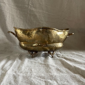 May include: A hammered brass bowl with a wavy rim and two small handles. The bowl rests on three ornate feet. The brass has a warm, golden hue, and the bowl appears to be vintage.