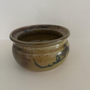 May include: A small, round, handmade ceramic bowl with a wide, flat rim. The exterior features a mottled tan and brown glaze with a dark, hand-painted swirling brushstroke design. The interior is smooth and glazed in a deep, earthy brown tone.