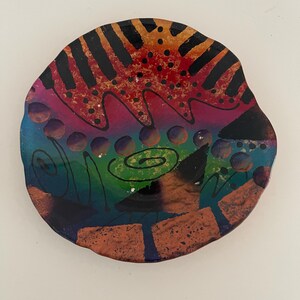 May include: A round, fused glass plate featuring a vibrant, multi-colored abstract design. The surface displays a gradient of red, orange, purple, green, and blue, accented with black painted lines, dots, spirals, and textured copper-colored shapes.