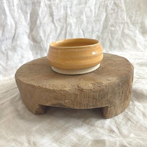 May include: A small, handmade ceramic bowl with a light orange glaze sits on a rustic, round wooden pedestal. The bowl has a simple, rounded shape and a textured surface. The pedestal is made of weathered wood.