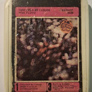 Peut inclure: Ancienne cassette 8 pistes de l'album "Obscured by Clouds" de Pink Floyd. Le boîtier en plastique blanc présente une étiquette rose et l'illustration de l'album. La liste des titres est visible en bas.