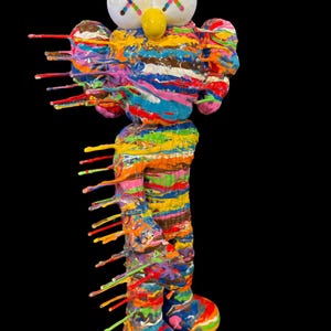 May include: A colorful, abstract sculpture of a cartoon figure with white eyes marked with an "X" and a yellow nose. The figure is covered in dripping paint in various colors, including blue, red, yellow, and pink. The background is black.