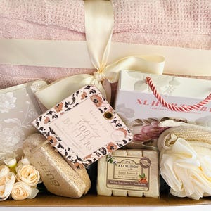 May include: A gift basket filled with self-care items. It includes a pink towel, a floral-patterned cosmetic bag, a box of foot buffers, a loofah, a bar of soap, and a small bouquet of flowers. The basket is tied with a cream-colored ribbon.