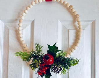 May include: A round Christmas wreath made of light-colored wooden beads. The wreath is decorated with green pine needles, red berries, a red apple, and a pine cone. A red ribbon is used for hanging.