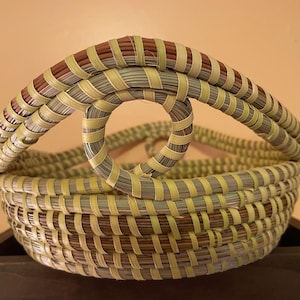 May include: A round woven basket featuring a coiled construction with alternating bands of light green, tan, and dark brown fibers. The basket includes a prominent arched handle that connects to a central circular loop design on the top rim.