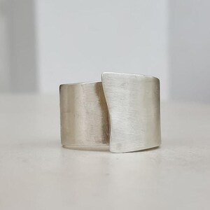 Può includere: Anello argentato dal design moderno e geometrico. L'anello presenta un'ampia fascia che avvolge il dito, con una sezione sovrapposta, creando una forma asimmetrica unica. La superficie ha una texture spazzolata.