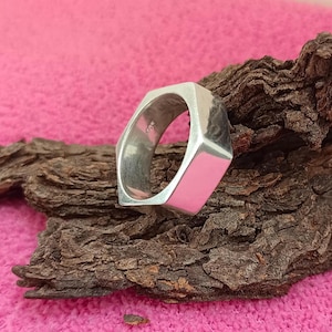 May include: A silver hexagonal ring, reflecting light. The ring is set against a backdrop of dark brown wood and a pink surface. The ring's design is geometric and modern, showcasing a polished finish.