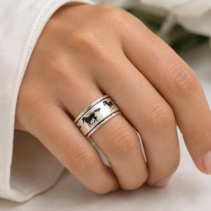 May include: A wide silver band ring featuring a repeating pattern of black horse silhouettes engraved around the exterior surface. The metal has a polished finish with darkened recessed details to highlight the equine figures.