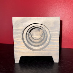 Puede incluir: Un bloque de madera gris con una serie de cortes circulares concéntricos. El bloque tiene forma cuadrada con pequeños pies en la base. El fondo es una pared roja lisa.