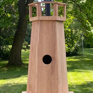 Puede incluir: Un comedero para pájaros en forma de faro de madera con una luz solar en la parte superior. El comedero para pájaros está hecho de madera de color marrón claro y presenta una abertura redonda. El diseño incluye una pequeña plataforma cuadrada y una barandilla decorativa.