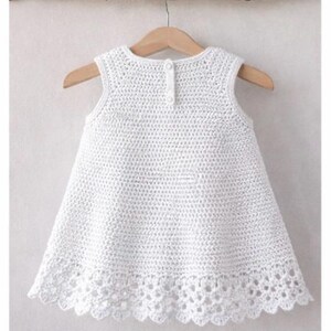May include: A sleeveless white crochet dress featuring a textured stitch pattern throughout the bodice and a decorative scalloped lace border at the hem. The garment includes a small button closure at the center of the upper back neckline.
