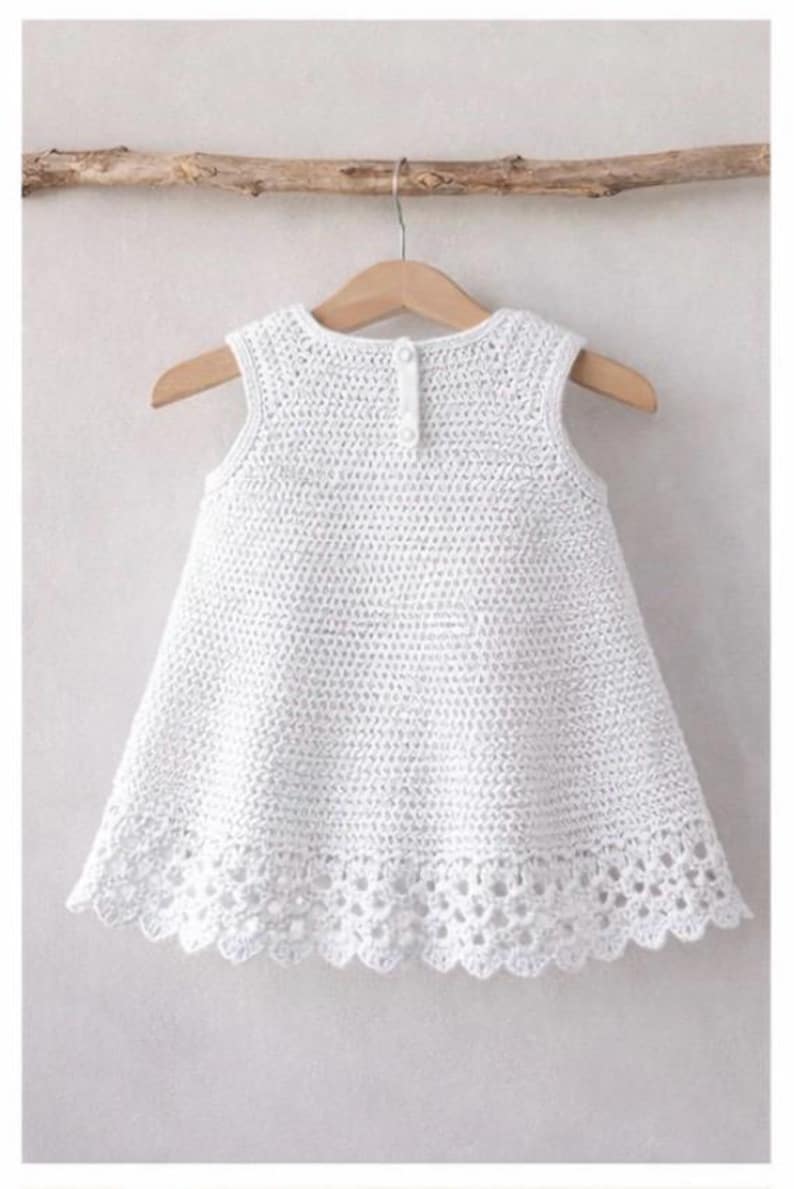 May include: A sleeveless white crochet dress featuring a textured stitch pattern throughout the bodice and a decorative scalloped lace border at the hem. The garment includes a small button closure at the center of the upper back neckline.