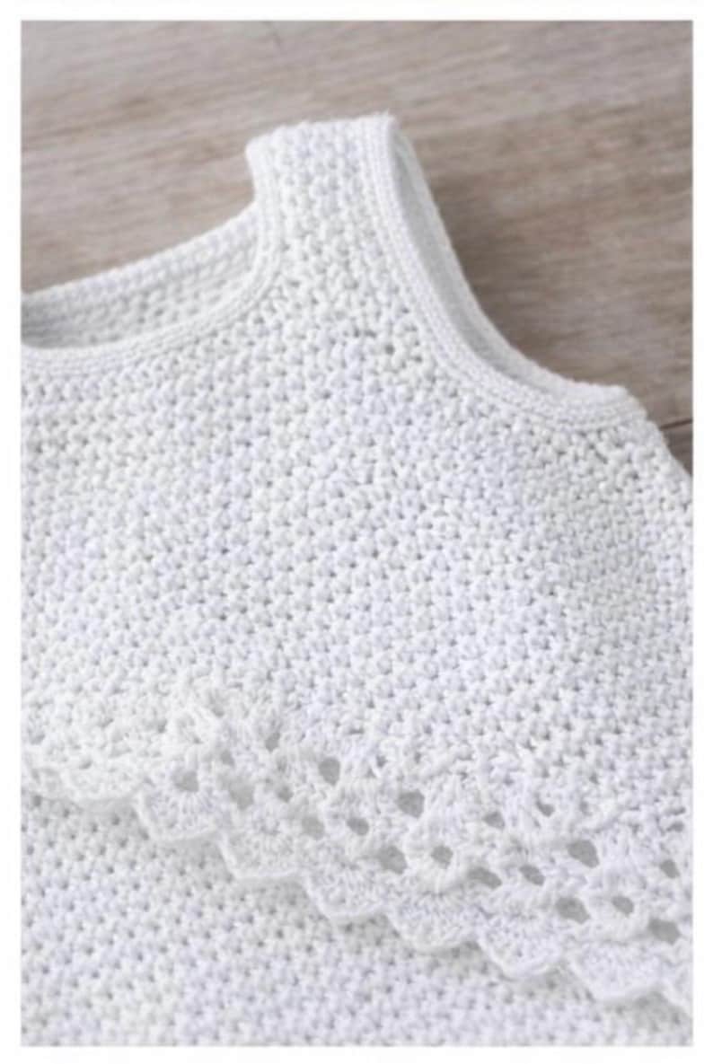 May include: A sleeveless white top featuring a dense crochet knit construction with a rounded neckline and a decorative scalloped edge detail that drapes across the chest, showcasing a uniform texture throughout the entire garment.
