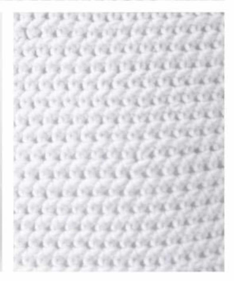 May include: A close-up view of a white textile featuring a consistent, repeating crochet stitch pattern. The surface displays a dense, textured arrangement of interconnected loops, creating a uniform and tactile appearance across the entire fabric.