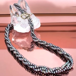 May include: A multi-strand necklace featuring dark gray pearls. The necklace is secured with a gold-colored clasp. The pearls are arranged in a twisted pattern, creating a textured appearance. The necklace is displayed on a reflective pink surface.