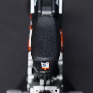 May include: A close-up shot of a black and orange motorcycle seat. The seat has a textured surface and is mounted on a silver frame. White accents are visible on the sides. The image is taken against a dark background.