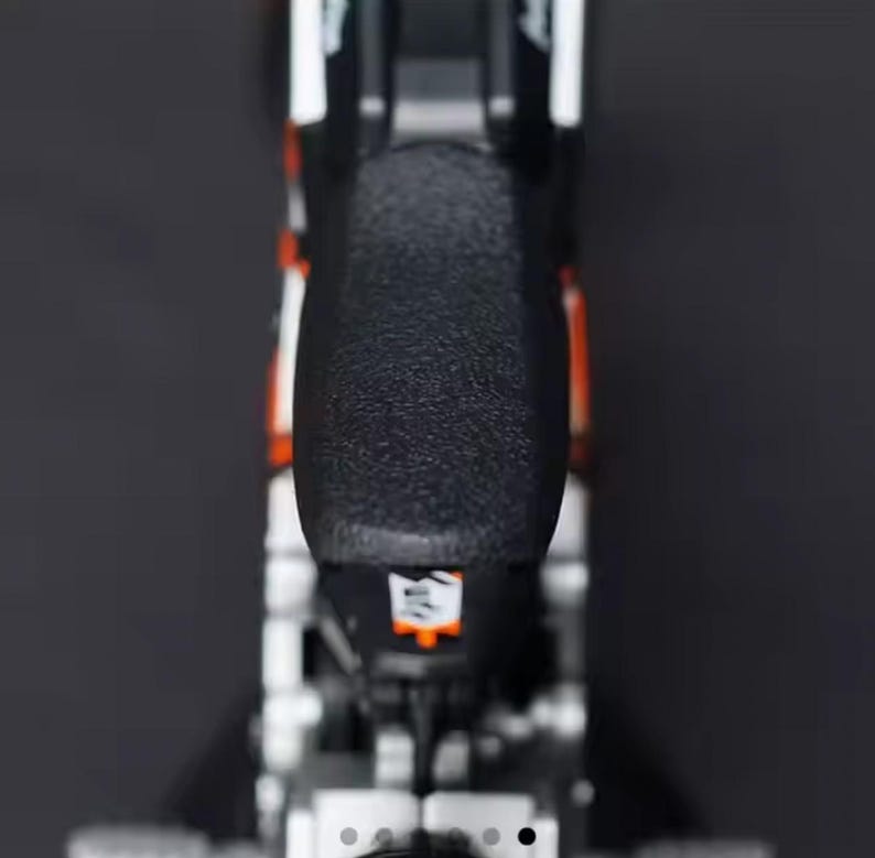 May include: A close-up shot of a black and orange motorcycle seat. The seat has a textured surface and is mounted on a silver frame. White accents are visible on the sides. The image is taken against a dark background.