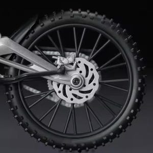 May include: Close-up of a black motorcycle wheel with a knobby tire. The wheel has silver spokes and a silver disc brake. The tire is thick and has a textured surface. The motorcycle's frame is visible.