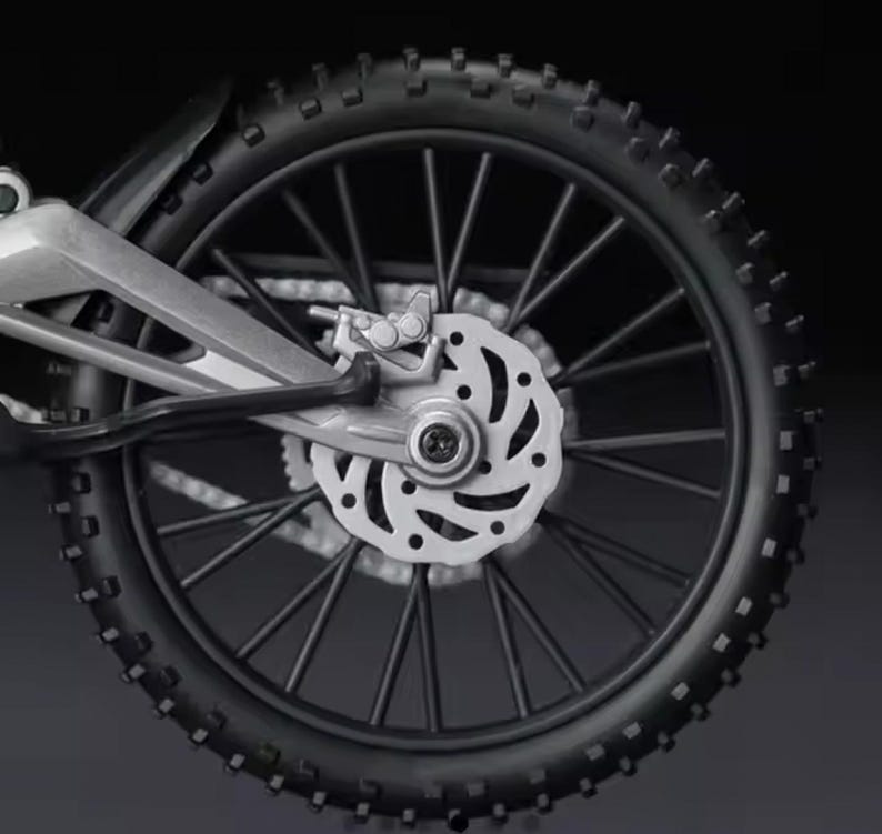 May include: Close-up of a black motorcycle wheel with a knobby tire. The wheel has silver spokes and a silver disc brake. The tire is thick and has a textured surface. The motorcycle's frame is visible.
