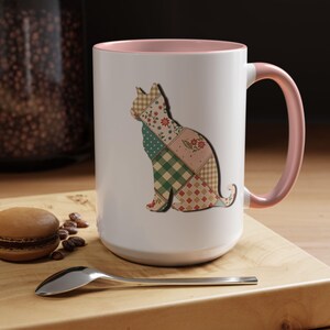 May include: White ceramic mug with a pink interior and handle. The mug features a patchwork cat silhouette in shades of green, pink, and tan. The mug sits on a wooden surface with a macaron, coffee beans, and a spoon.