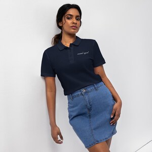 May include: A navy blue polo shirt with the text "created equal" embroidered in white. The shirt is paired with a denim skirt. The model is leaning against a white wall.