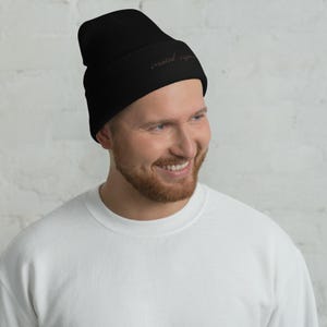 Op de afbeelding: Een zwarte beanie met de geborduurde tekst "created equal" in lichtbruin. De beanie is gemaakt van een zacht, geribbeld gebreid materiaal. De persoon op de foto draagt een witte trui.