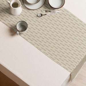 Puede incluir: Camino de mesa beige con un patrón repetido de texto pequeño. Se coloca sobre un mantel blanco, con una jarra blanca, una taza, platos, cuencos y cubiertos visibles. El camino de mesa añade un toque decorativo a la mesa.