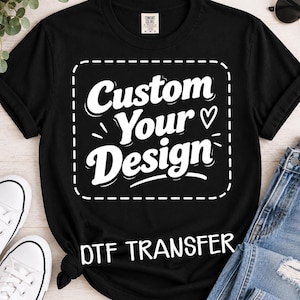 May include: A black short-sleeve t-shirt featuring a white graphic print with the text Custom Your Design inside a dashed square border, with the additional text DTF TRANSFER printed below the square, displayed on a flat surface.