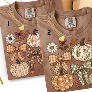May include: Two brown t-shirts featuring a fall-themed graphic print. The design includes pumpkins with floral and plaid patterns, checkered bows, acorns, and daisies. Each shirt has a Comfort Colors label at the neck and a gold heart necklace.
