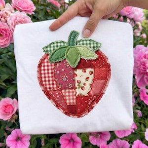 May include: A white t-shirt featuring a large, appliqued strawberry design. The strawberry is composed of various red fabric patches, including gingham and floral patterns. Green gingham fabric forms the stem and leaves. The design is held by a hand.