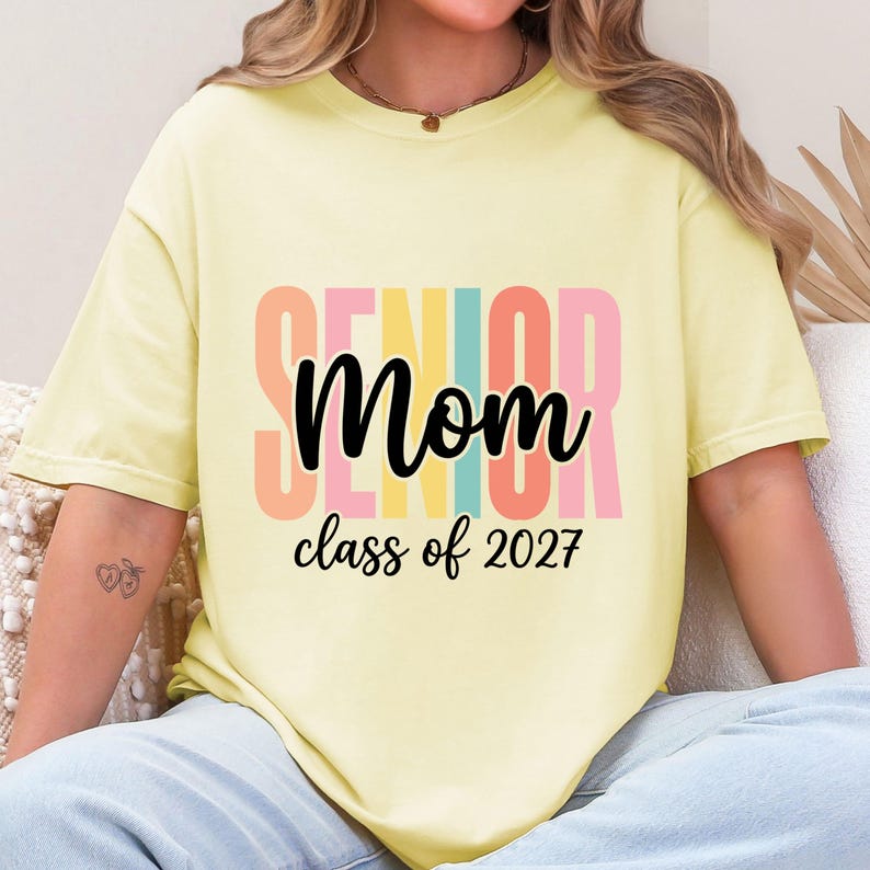 May include: A pale yellow short-sleeve t-shirt featuring the word SENIOR in large, vertical, multi-colored block letters behind the black cursive text Mom and the black printed text class of 2027 centered on the front of the garment.