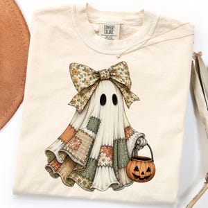 May include: A cream-colored t-shirt featuring a graphic of a ghost wearing a large floral bow and a patchwork-style sheet. The ghost holds a small orange jack-o'-lantern bucket. The shirt has a Comfort Colors brand label at the inner neckline.