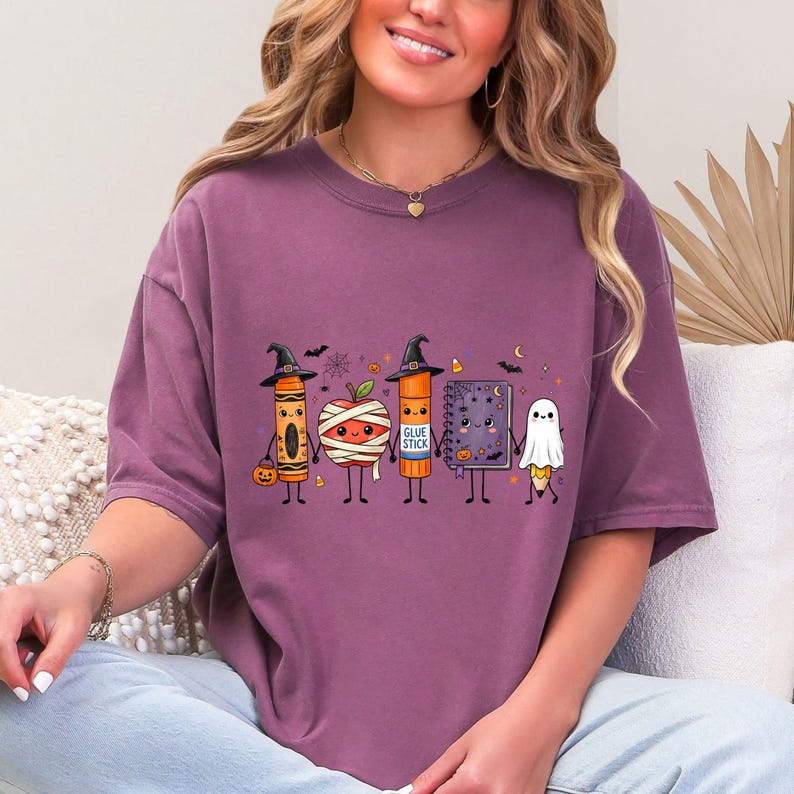 May include: A mauve short-sleeve t-shirt featuring a graphic of five cute Halloween-themed school supplies holding hands: a crayon in a witch hat, a mummy apple, a glue stick in a witch hat, a purple notebook, and a pencil dressed as a ghost.