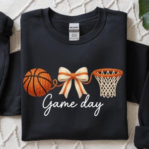 May include: Black sweatshirt with an orange basketball, bow, and net design. The words "Game day" are written in white script below the basketball graphic. The sweatshirt is a Gildan brand.