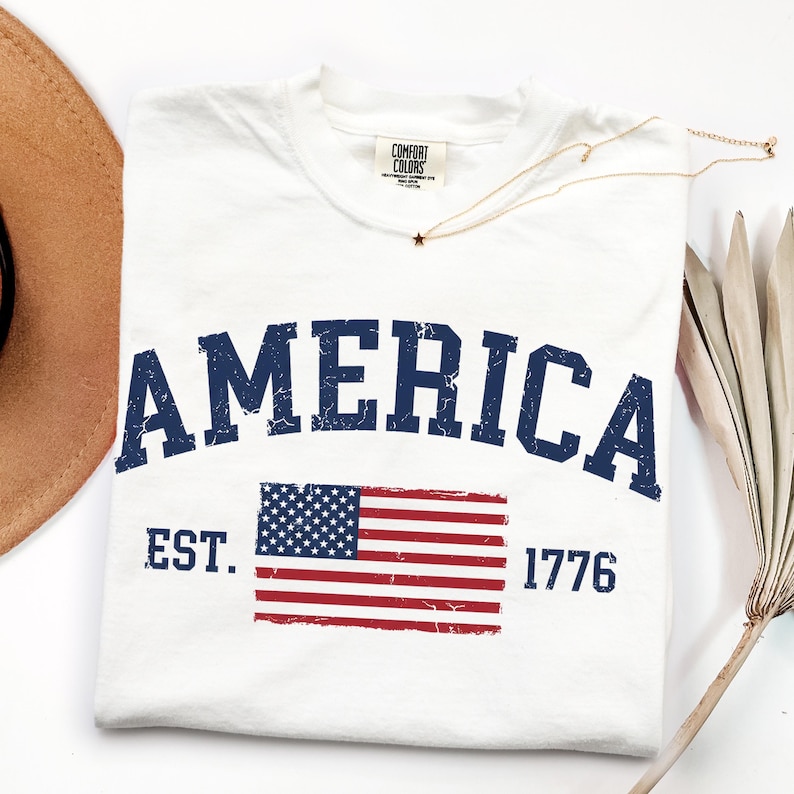 May include: A white short-sleeved t-shirt featuring the word AMERICA in large, distressed navy blue letters arched across the chest. Below the text is a graphic of the American flag with the text EST. 1776 in navy blue on either side.